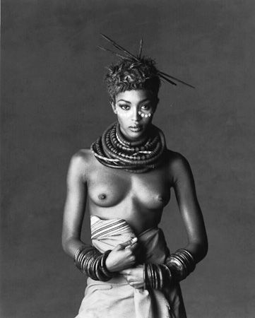 A modelo negra Naomi Campbell, posa com adereços da cultura africana. Usa um turbante no cabelo, colares volumosos que cobrem toda a extensão do seu pescoço e  usa pulseiras que cobrem seus pulsos. Seus seios estão à mostra, usa apenas uma saia. À foto recorta à extensão da cabeça até os joelhos, não mostrando as pernas e pés.