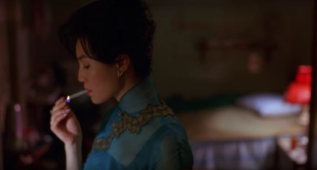 This is a screen still from In The Mood for Love. A woman is framed in medium close-up. A cigarette is between her lips and she's lifting up a match to light it,