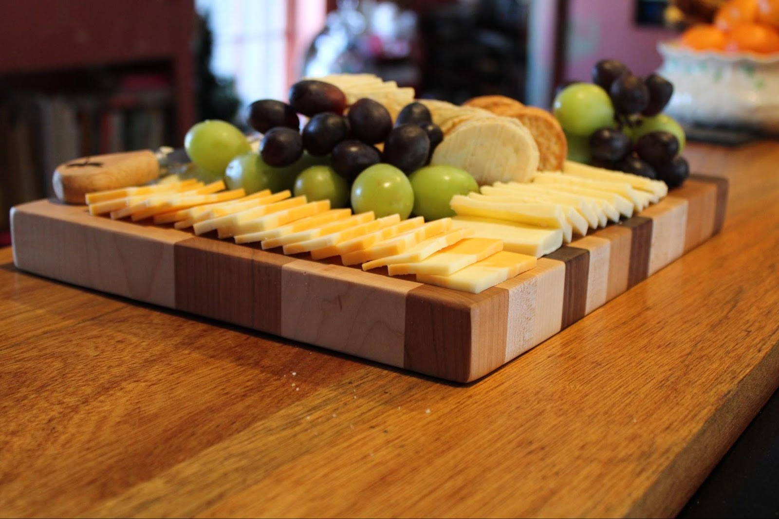 handmade maple cherry and walnut charcuterie board topped with cheeses, crackers, and grapes