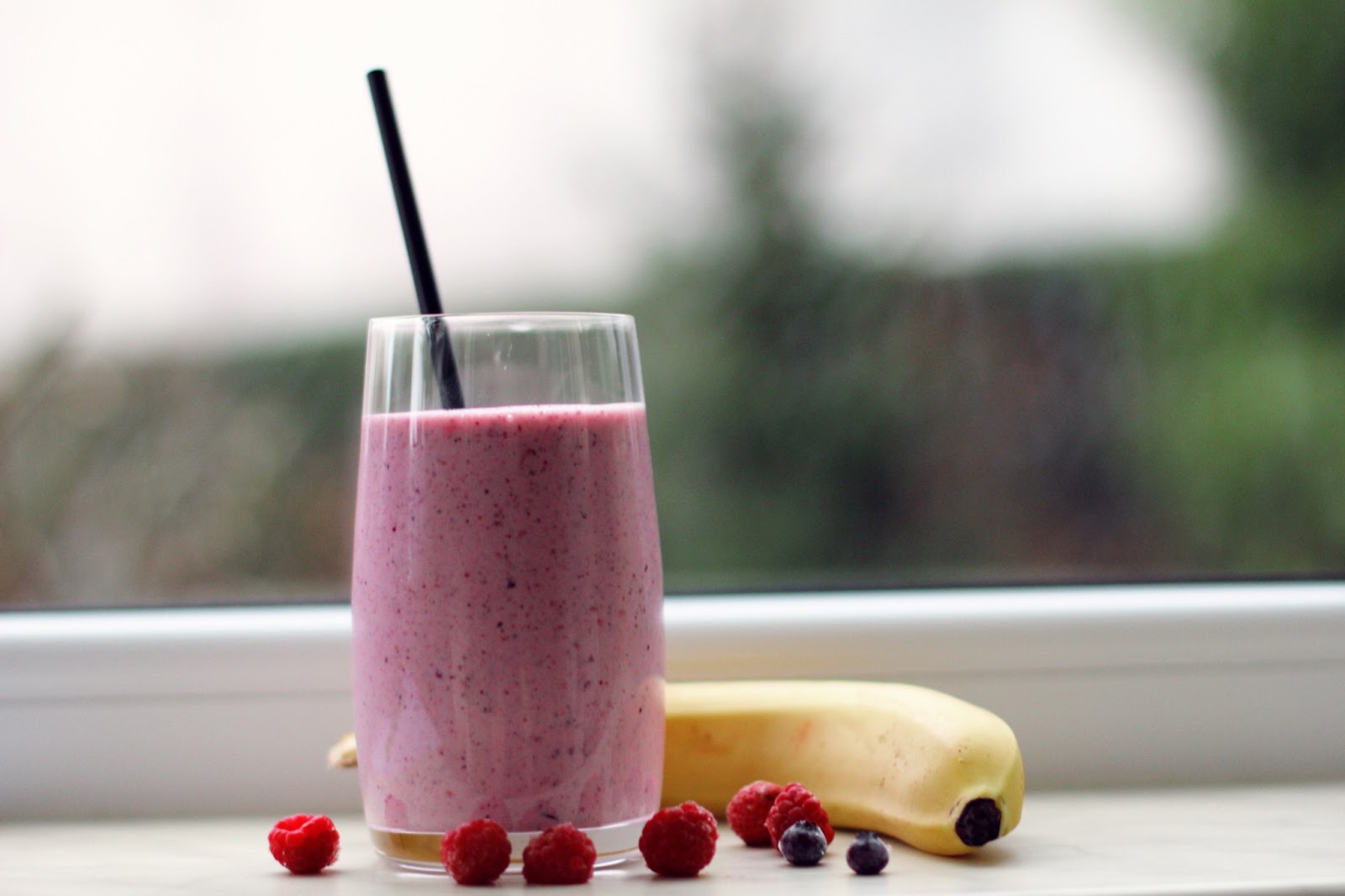 A pink fruit smoothie is pictured in a glass with a straw, on a counter next to raspberries, blueberries, and a banana