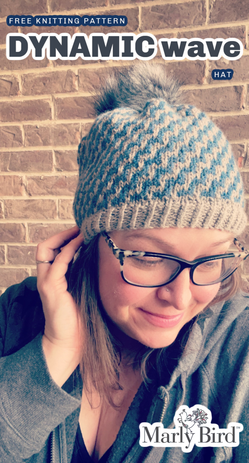 A cheerful person in glasses sporting a cozy mosaic knit beanie smiles for a selfie against a brick wall background. Dynamic Wave Mosaic Knit Hat Pattern by Marly Bird in blue and light gray color.