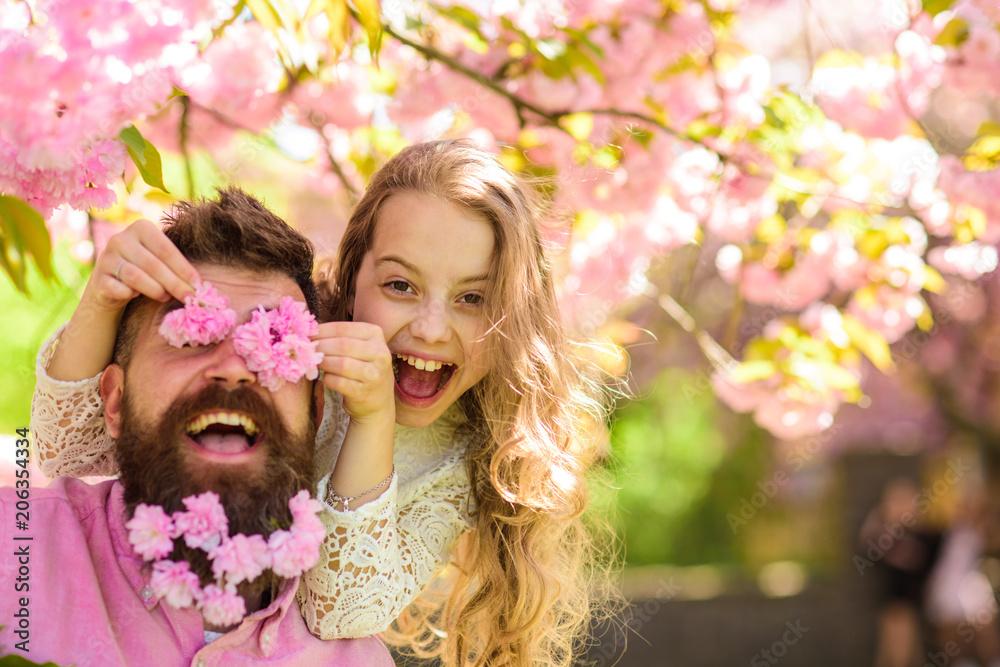 Child and man with tender pink flowers in beard. Girl with dad near sakura flowers on spring day. Father and daughter on happy face play with flowers as glasses, sakura background. Family time concept