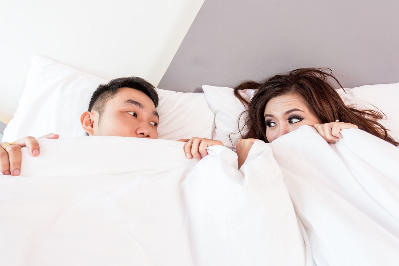 A young couple lying in bed looking at each other while under a blanket.