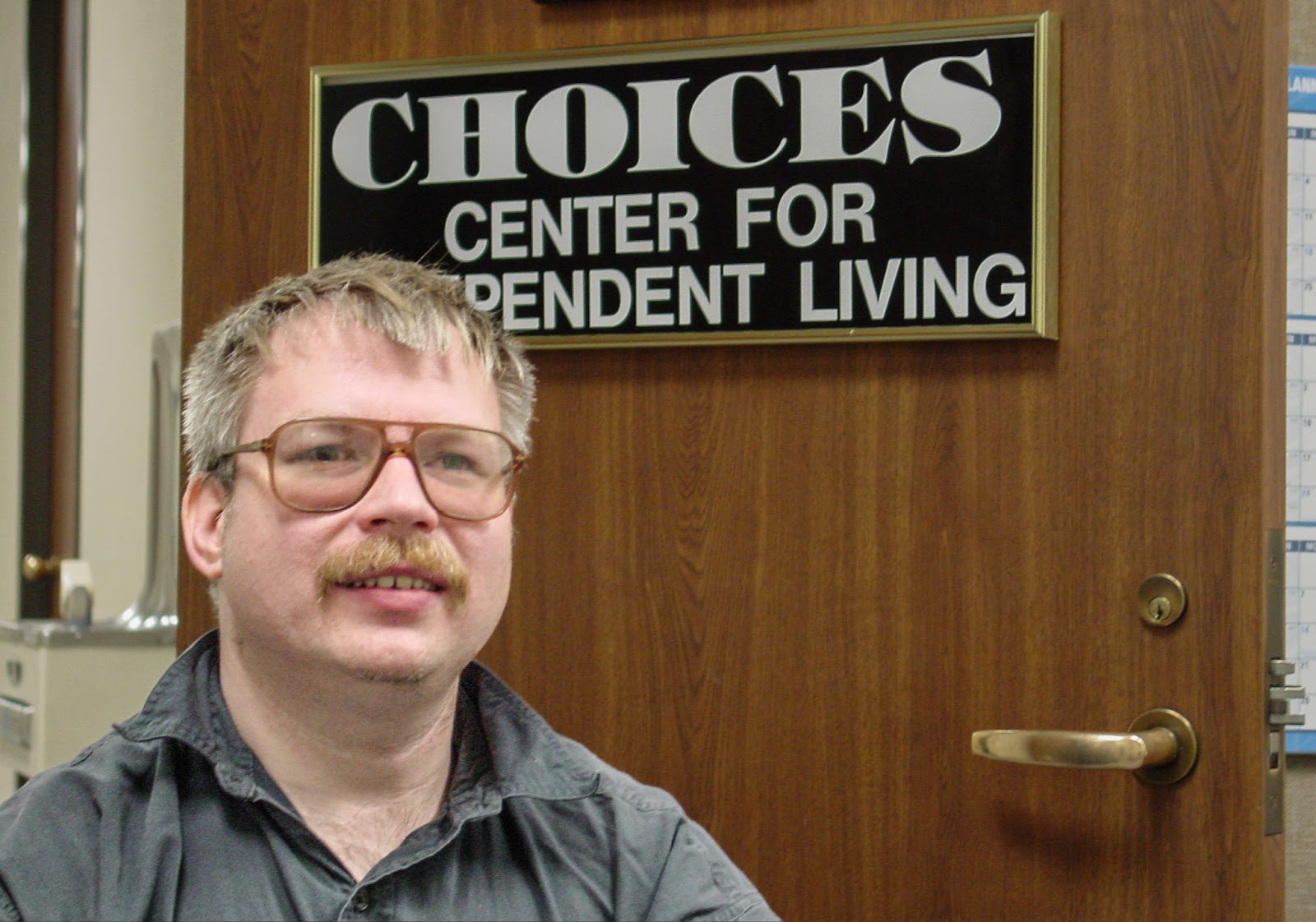 Man with mustache and glasses in front of a sign that reads: CHOICES CENTER FOR INDEPENDENT LIVING.