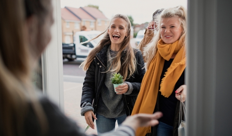 Three young women are standing at their neighbor's front door, carrying gifts and smiling. They're coming to meet their new neighbor.