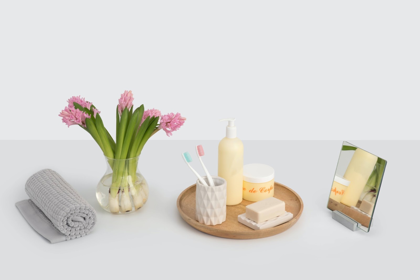 Foto di alcuni prodotti di bellezza e di igiene orale disposti su una superficie bianca con un vaso di fiori, un asciugamano e uno specchio