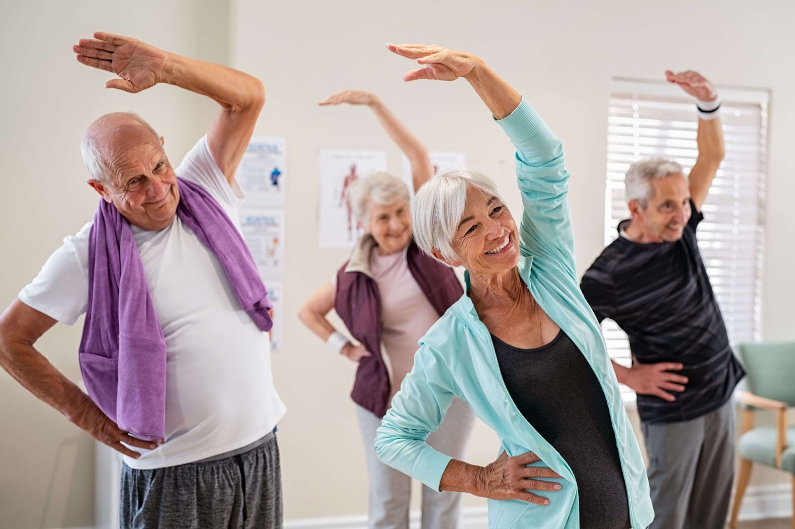 A group of seniors in an exercise class smiling and stretching with one hand over their heads.