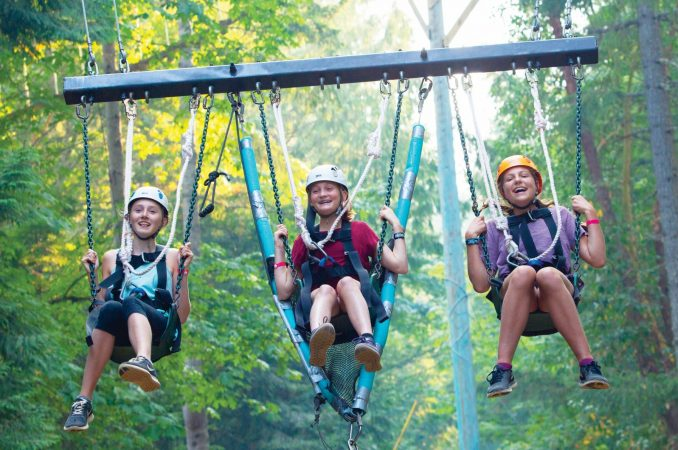 Three children soaring through the woods on a swing, experiencing an exhilarating adventure at Camp Qwanoes.