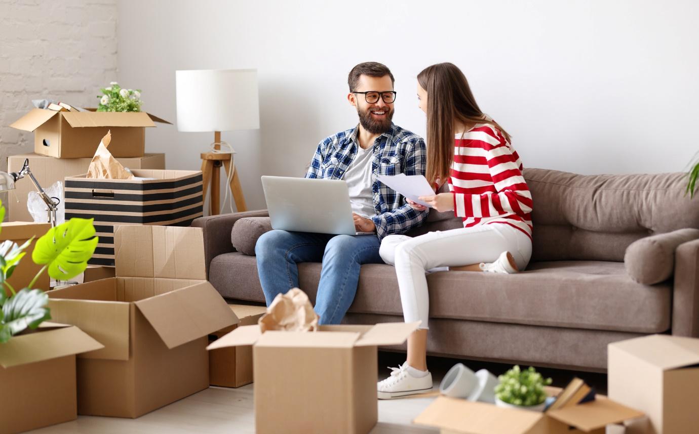 couple in new home sitting on sofa surrounded by boxes checking a laptop