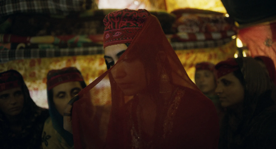 A scene from The Shepherdess and the Seven Songs, featuring Laila dressed in red with a red veil, surrounded by other women in a dimly lit tent.