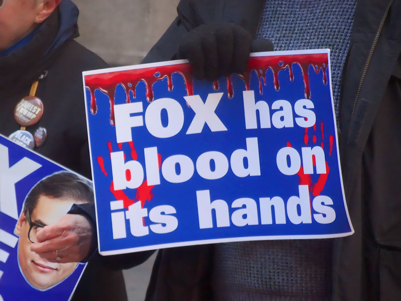 A man wearing a blue sweater, black jacket, and black gloves with his head cropped out holds a blue sign with an image of blood dripping down the top and a red handprint on both sides. Overlaid are white letters that read “FOX has blood on its hands.”