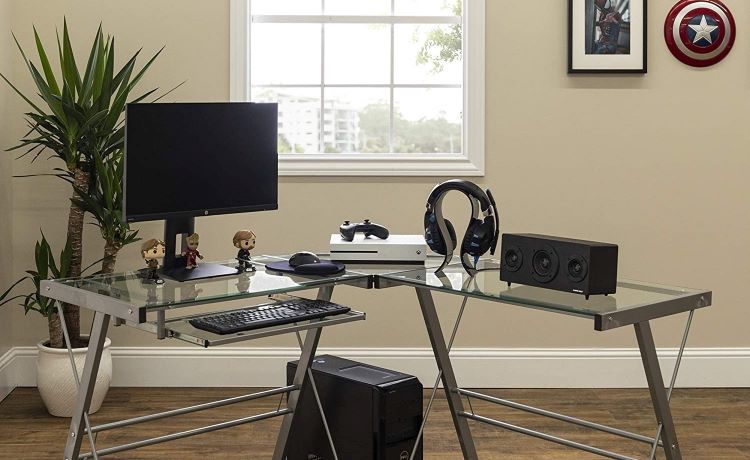  A glass and metal, corner office desk setup in a home office. There’s a large monitor, keyboard, speakers, and headphones on the desk, as well as a gaming station and some bobble heads. 