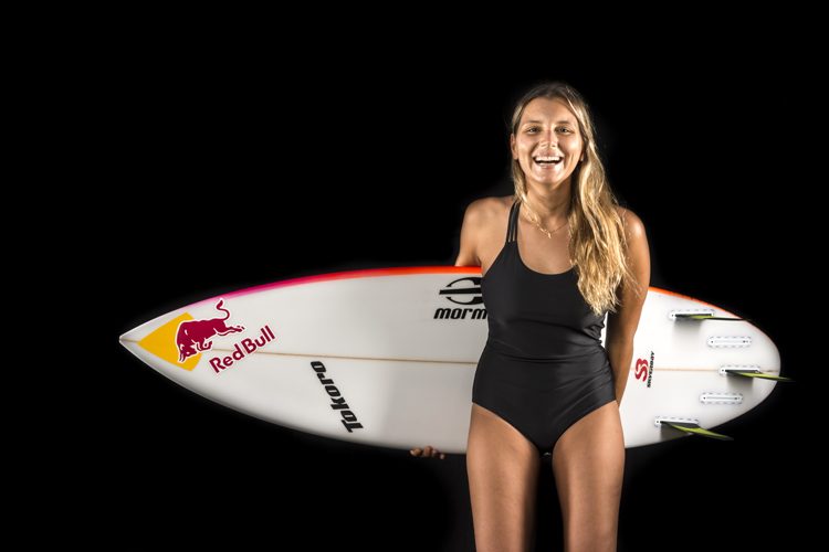 #DescriçãoDaImagem: mulher branca, loira, está de pé sorrindo, vestindo um maiô preto e segura uma prancha de surf branca com escritos em preto e vermelho. Foto: Surfer Today.