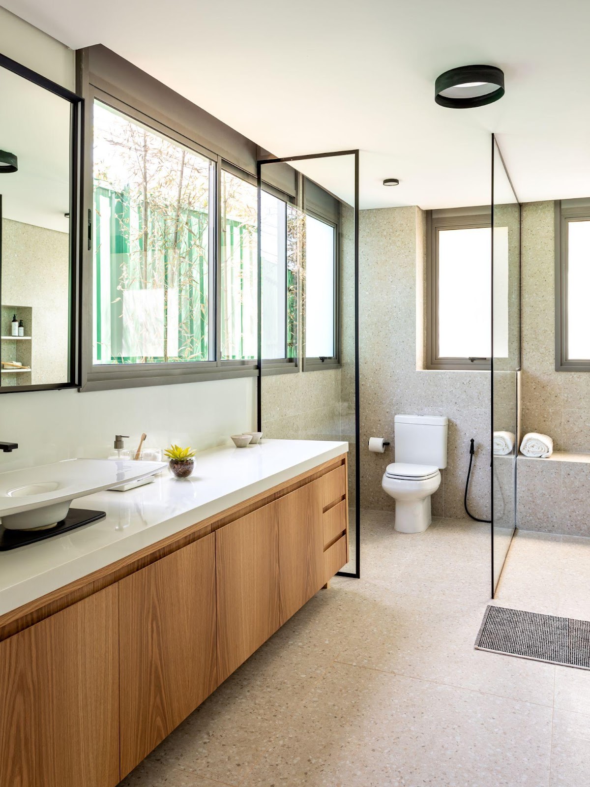 Banheiro com piso e parede revestidos de granilite, box de vidro, gabinete grande de madeira com bancada e cuba branca.