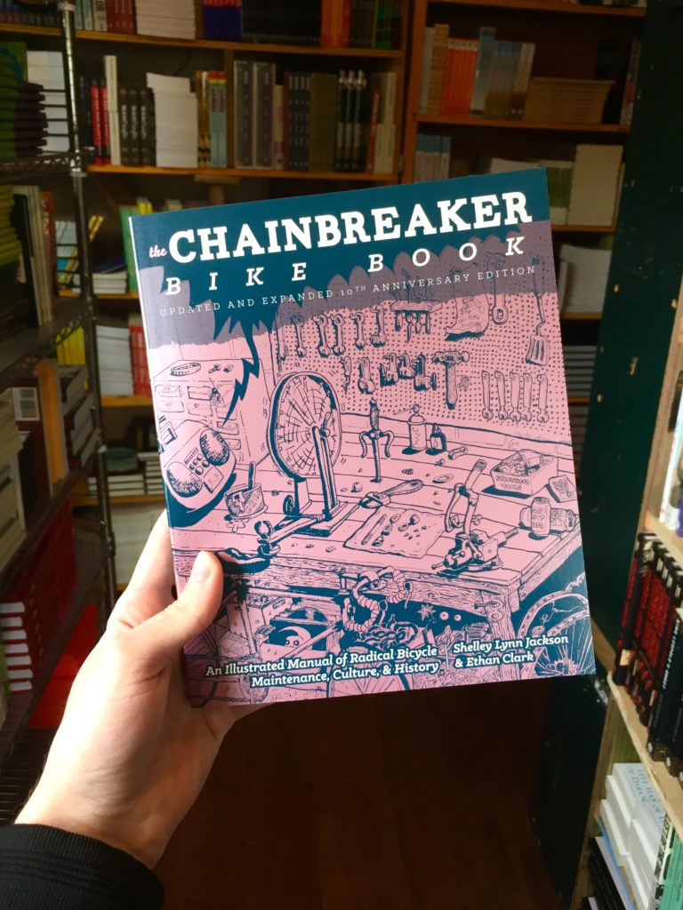 Photo of a hand holding the new edition of Chainbreaker Bike Book with bookshelves in the background