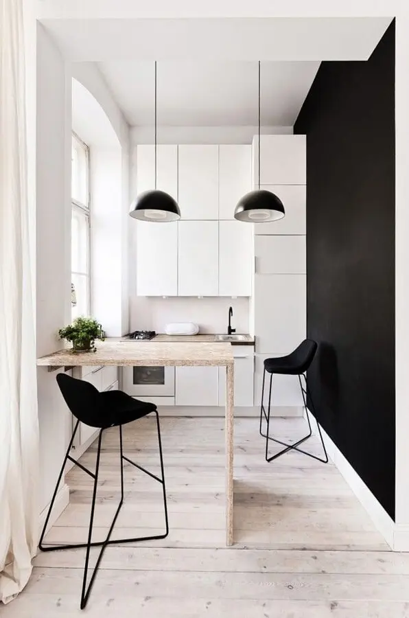 Cozinha com armários brancos e parede lateral preta. Na frente da pia há uma bancada de madeira clara com dois bancos pretos. Um pequeno vaso de planta decorativo.
