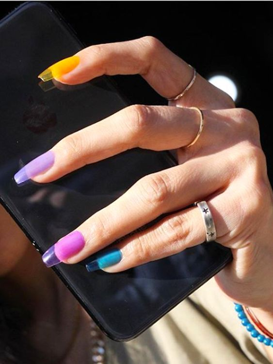 #DescriçãoDaImagem: uma mão tem anéis prateados em três dedos, segura um celular preto e as unhas estão pintadas de amarelo, lilás, roxo e azul cada uma. Foto: Pinterest.