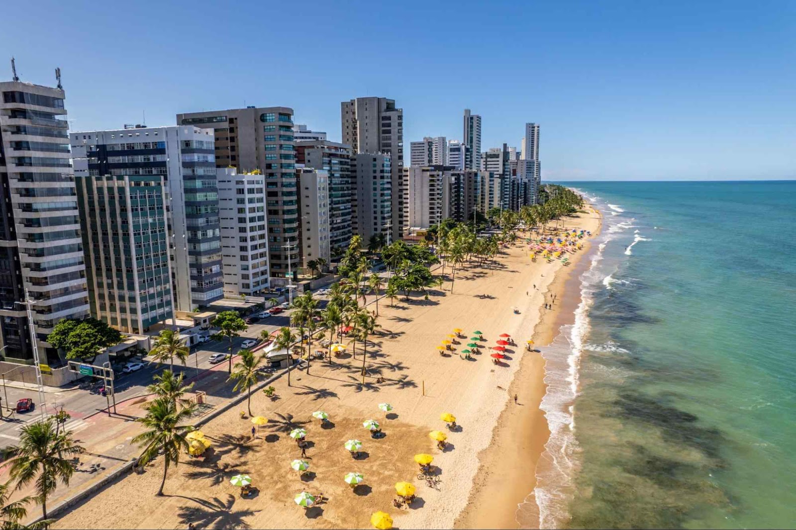 Praia de Boa Viagem, Recife. A longa faixa de areia tem alguns guarda-sóis coloridos e poucos coqueiros. Ela é delimitada pelo mar esverdeado de um lado e por uma fileira de prédios altos do outro.