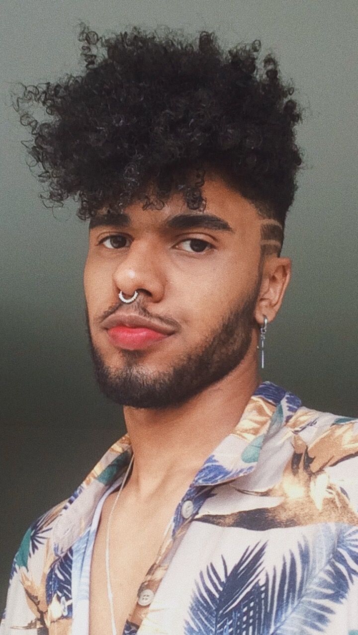 Um homem negro de cabelo cacheado cortado no estilo “pixie” aparece na imagem. Ele veste uma camisa com estampas tropicais.