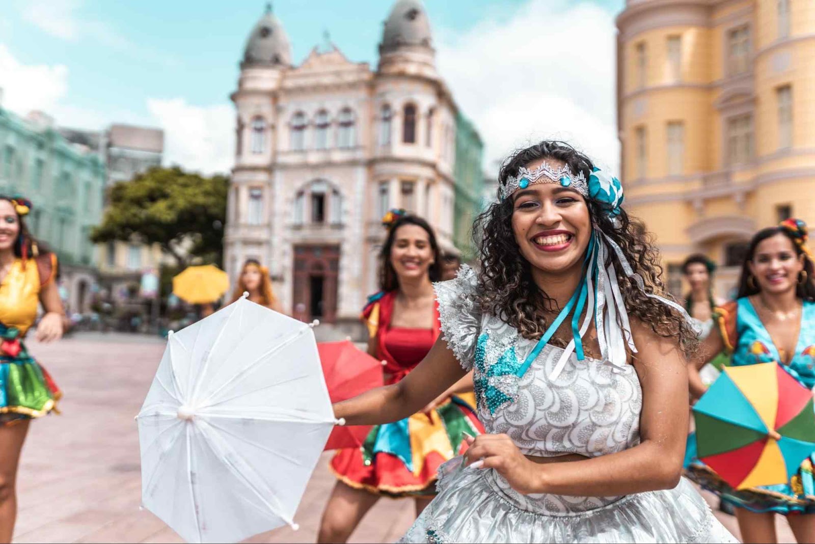 Passistas caracterizadas com trajes tradicionais do carnaval recifense. Elas dançam frevo em frente às construções históricas do Recife Antigo.