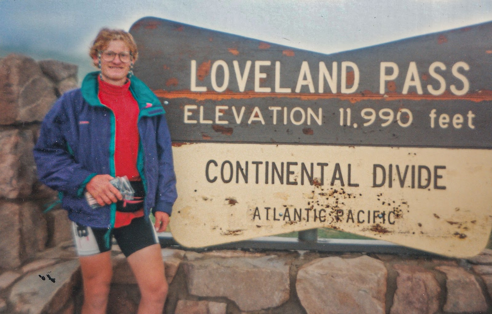 Man stands in coat with biking shorts by a sign that reads: LOVELAND PASS ELEVATION 11,990. CONTINENTAL DIVIDE ATLANTIC, PACIFIC