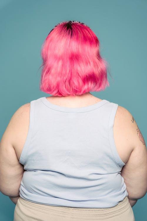 Back view of young plus size female in tank top with pink dyed hair standing against blue background
