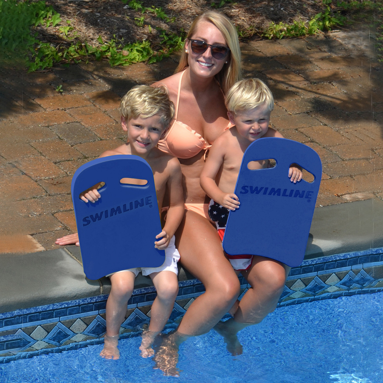 mother with two small boys sitting on the side of a pool and olding swim kickboards 