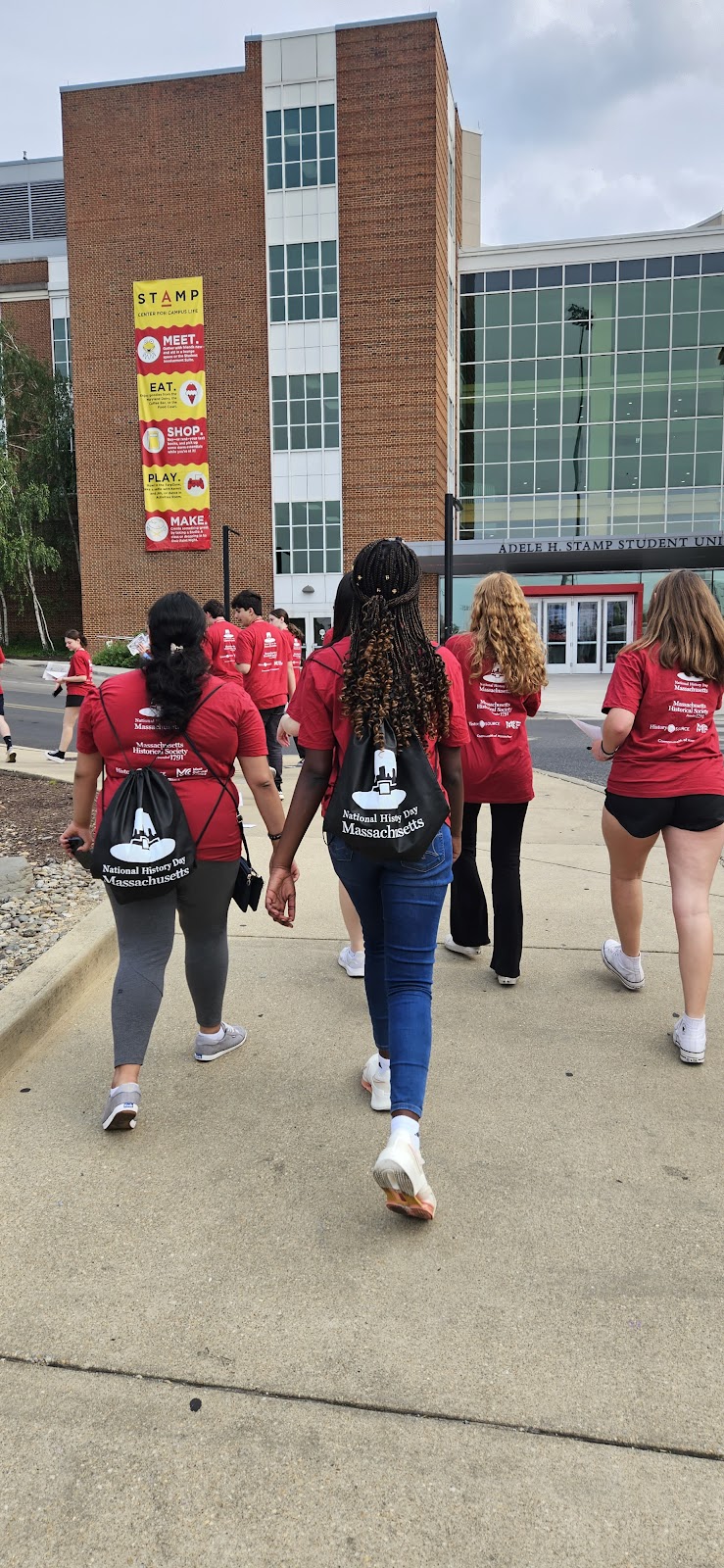 Image shows the backs of several students walking towards a building. Students are wearing red t-shirts and black drawstring bags with a white MHS History day logo imprinted.