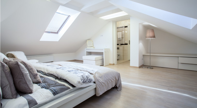A modern bedroom with unique gabled ceilings and skylights. The walls are painted white and the bed and other furniture are light-colored.