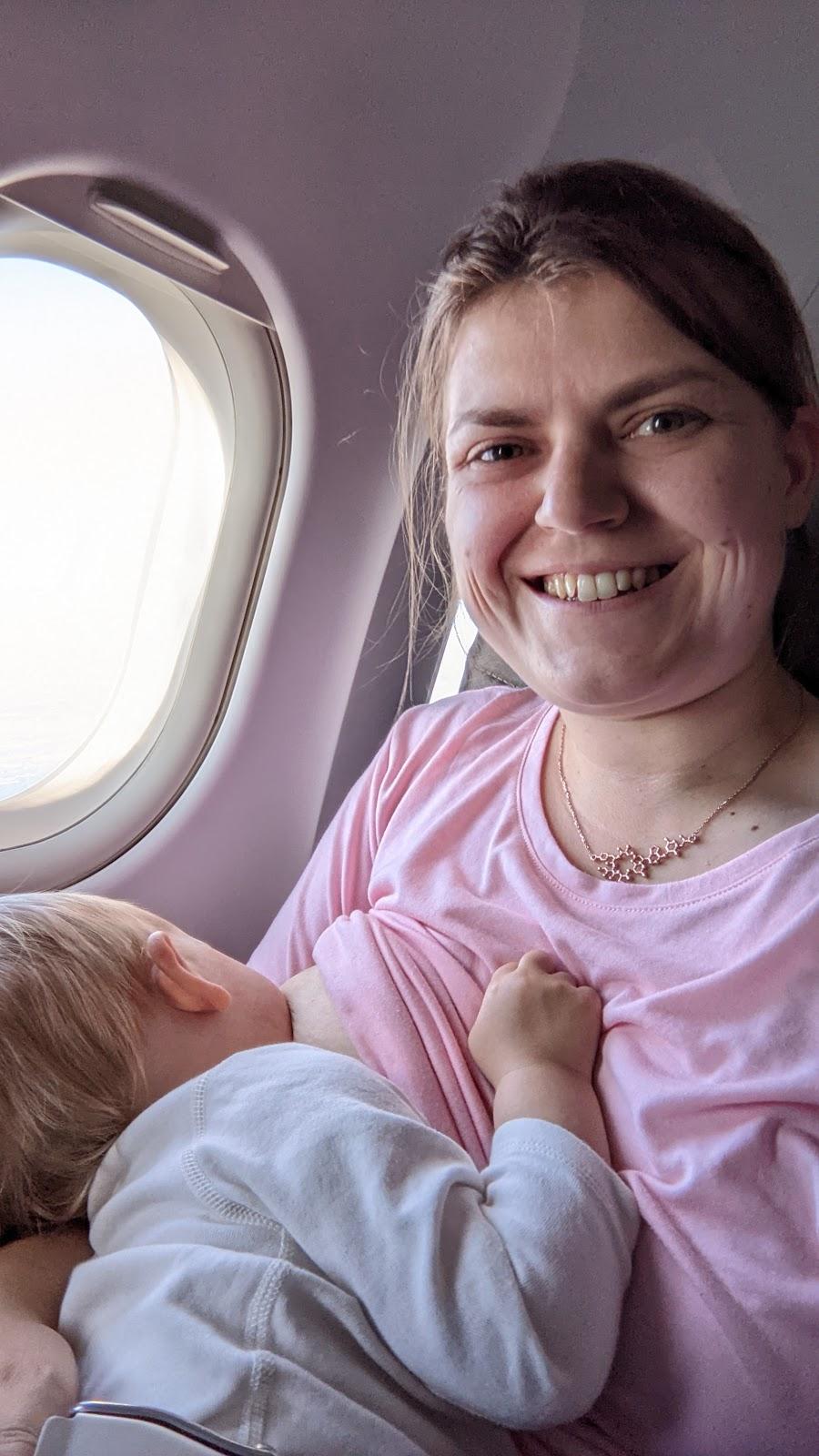 Fiona is breastfeeding her daughter while on a plane. Her daughter is laid across her lap with a plane seatbelt buckled around her. Fiona is wearing a pink t-shirt and a necklace in the shape of the chemical oxytocin.