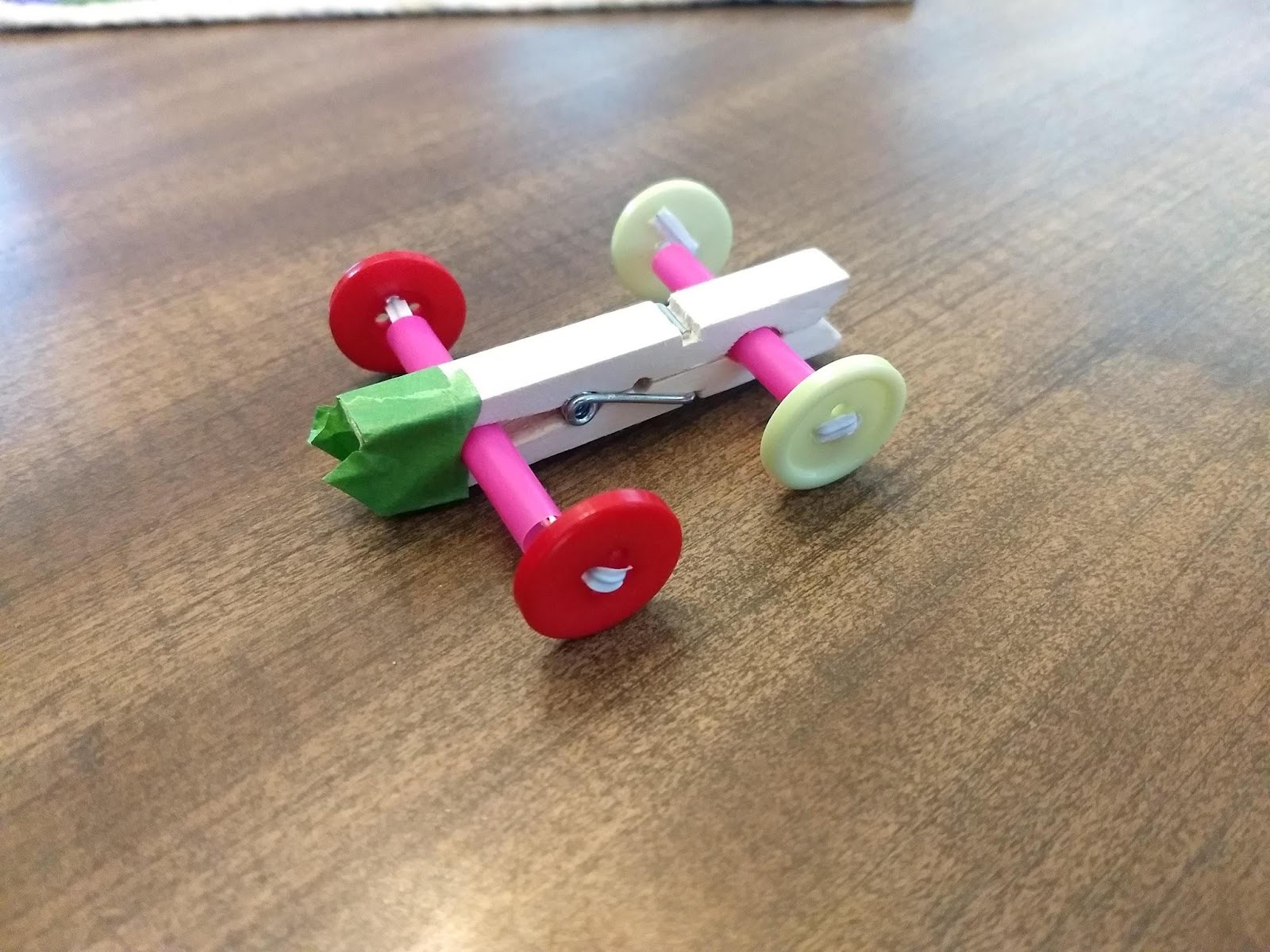 A simple machine clothespin car sits on a wooden table with red and green equally sized buttons as wheels with bread ties as axles.