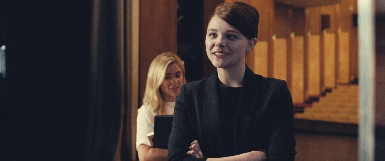 Chloe Grace Moretz as Jo-Ann playing Sigrid in 'Clouds of Sils Maria' (2014). Jo-Ann, dressed in black with her hair scraped back, looks smug, standing with her arms folded across her chest.