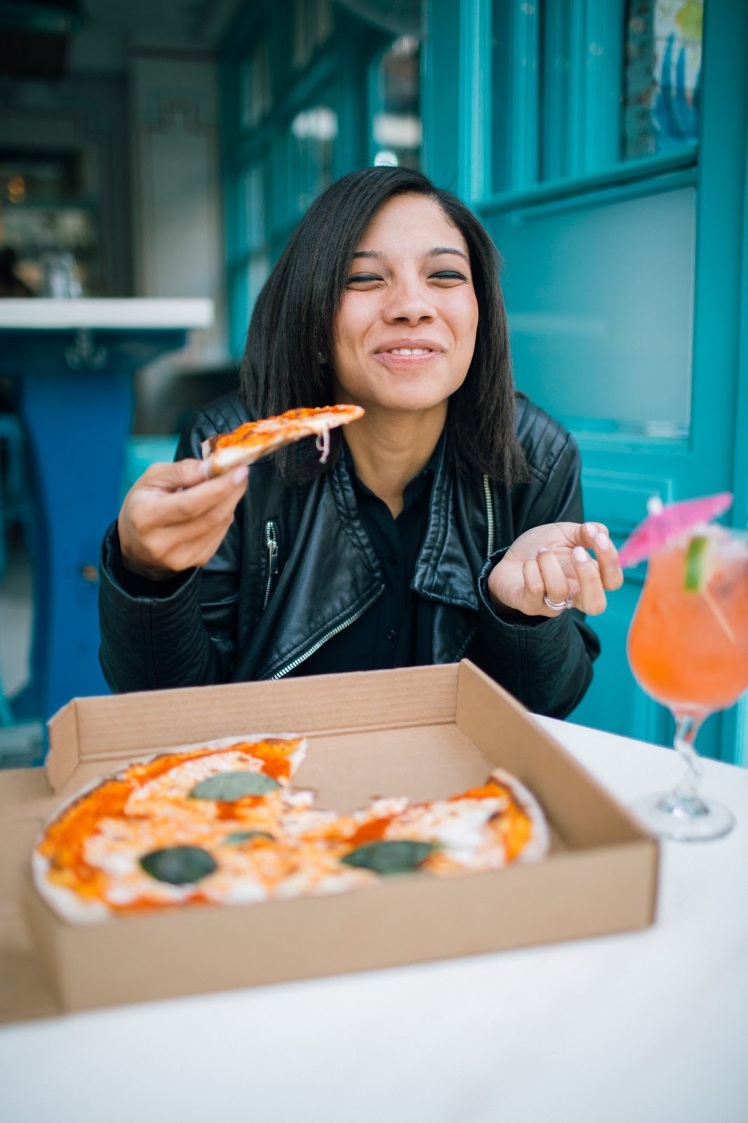 A woman smiles after taking a bite of a piece of pizza topped with cheese and basil leaves.