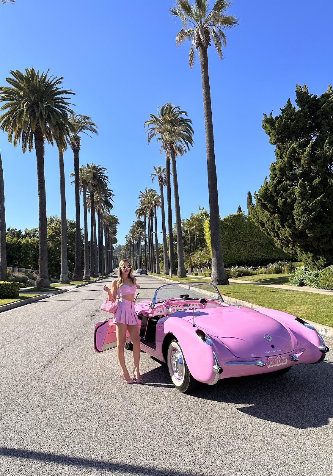 Margot Robbie standing beside a pink convertible in a palm-tree-lined Hollywood street.