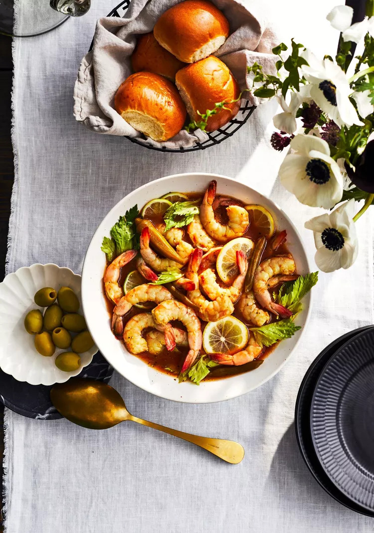 A beautifully set table with a bowl of lemon garlic shrimp garnished with celery leaves, a basket of freshly baked rolls, green olives, and a golden spoon, creating an inviting atmosphere for a delightful seafood meal.