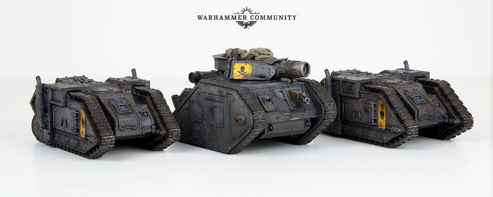 Three armoured fighting vehicles of the imperial army in a row. The two at the sides are Aurox transports, and the middle vehicle is a leman russ battle tank. They are painted in urban blue, with yellow unit marker details.