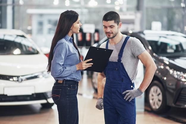 Foto gratuita hermosa joven está hablando con un guapo mecánico mientras repara un automóvil en concesionario