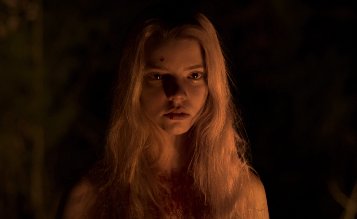 Still from The Witch. A close up of Thomasin, her naked body hidden by her long blonde hair. She is splattered with blood, illuminated only by a dim orange light.