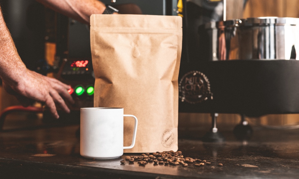 White coffee mug and brown, unbleached kraft paper coffee bag on counter with roaster working in background. 