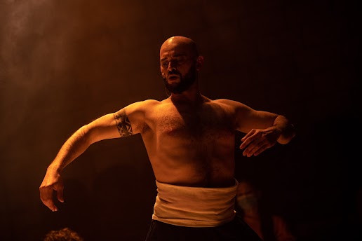 Photo Credit: Maria Baranova. Image Description: Christopher holds a strong gesture with his arms as an amber light bathes his body.