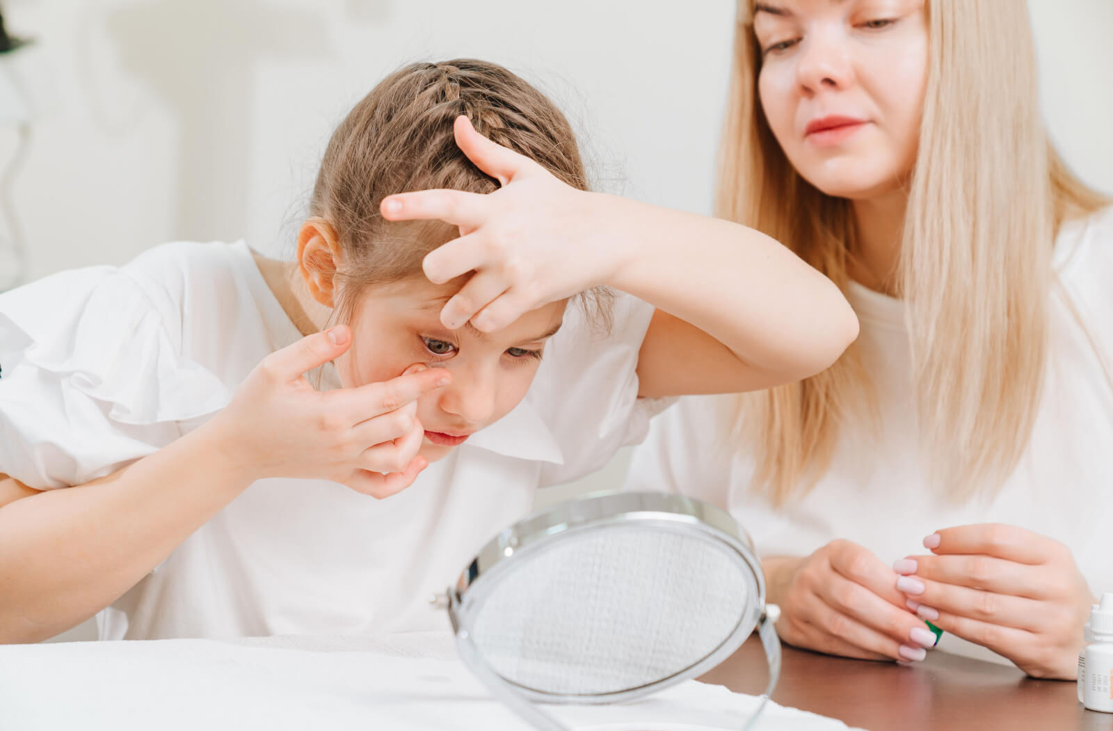 Young girl putting an effort to wear her first pair of contact lenses while being supervised by her mother.