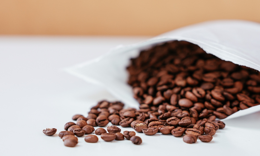 Roasted coffee beans tumbling out of a white, bleached kraft paper coffee pouch lying on its side on white table with sandstone colour background