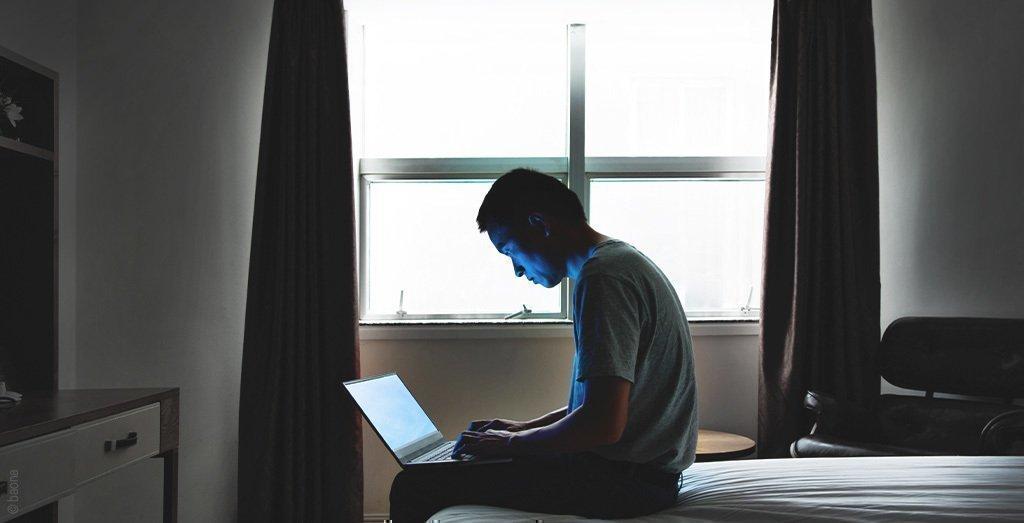 Foto: Un hombre está sentado en el borde de la cama en el dormitorio, con una computadora portátil en su regazo que le ilumina la cara.