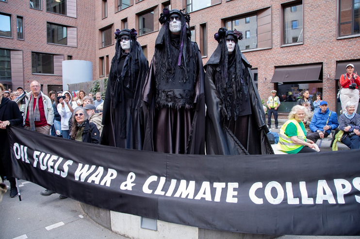 Three rebels in black robes stand behind a banner saying 'Oil fuels war and climate collapse'