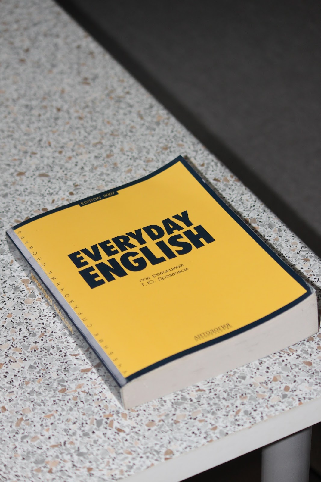 A imagem mostra um livro de capa amarela sob uma mesa de mármore. O livro possui o título “Everyday English”, traduzido como “Inglês diário”. 