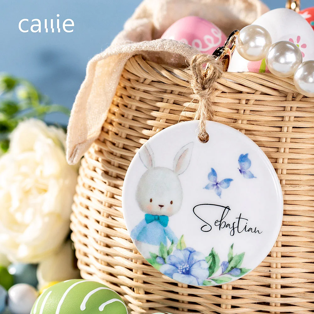 Étiquette personnalisée par un prénom, décorée d’un lapin de Pâques, de fleurs, de papillons et placée sur un panier de Pâques. 