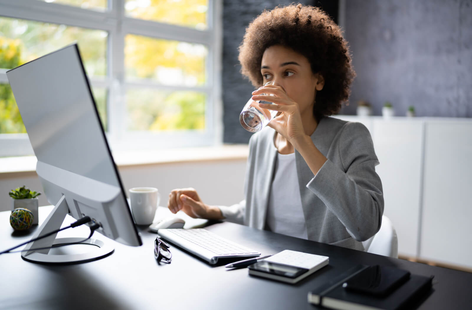 A professional-looking woman drinking water as she sits at a desk in front of a desktop computer.