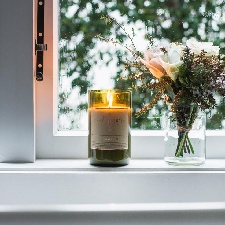 Cozy window scene with a lit candle in a green glass container and a bouquet of flowers in a glass jar, set against a rainy backdrop.