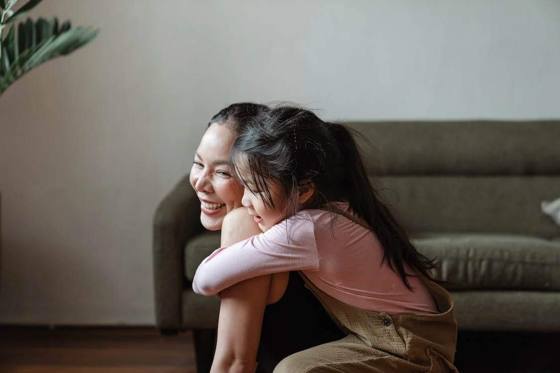 a little girl hugging her mum from behind, both smiling. 