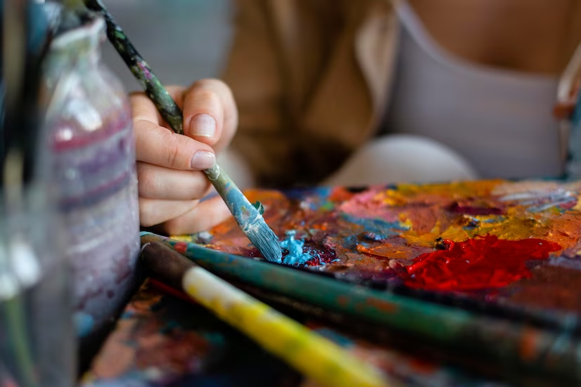 mão feminina segura um pincel de pintura sobre uma paleta de pintura antes de levar ao quadro que está produzindo